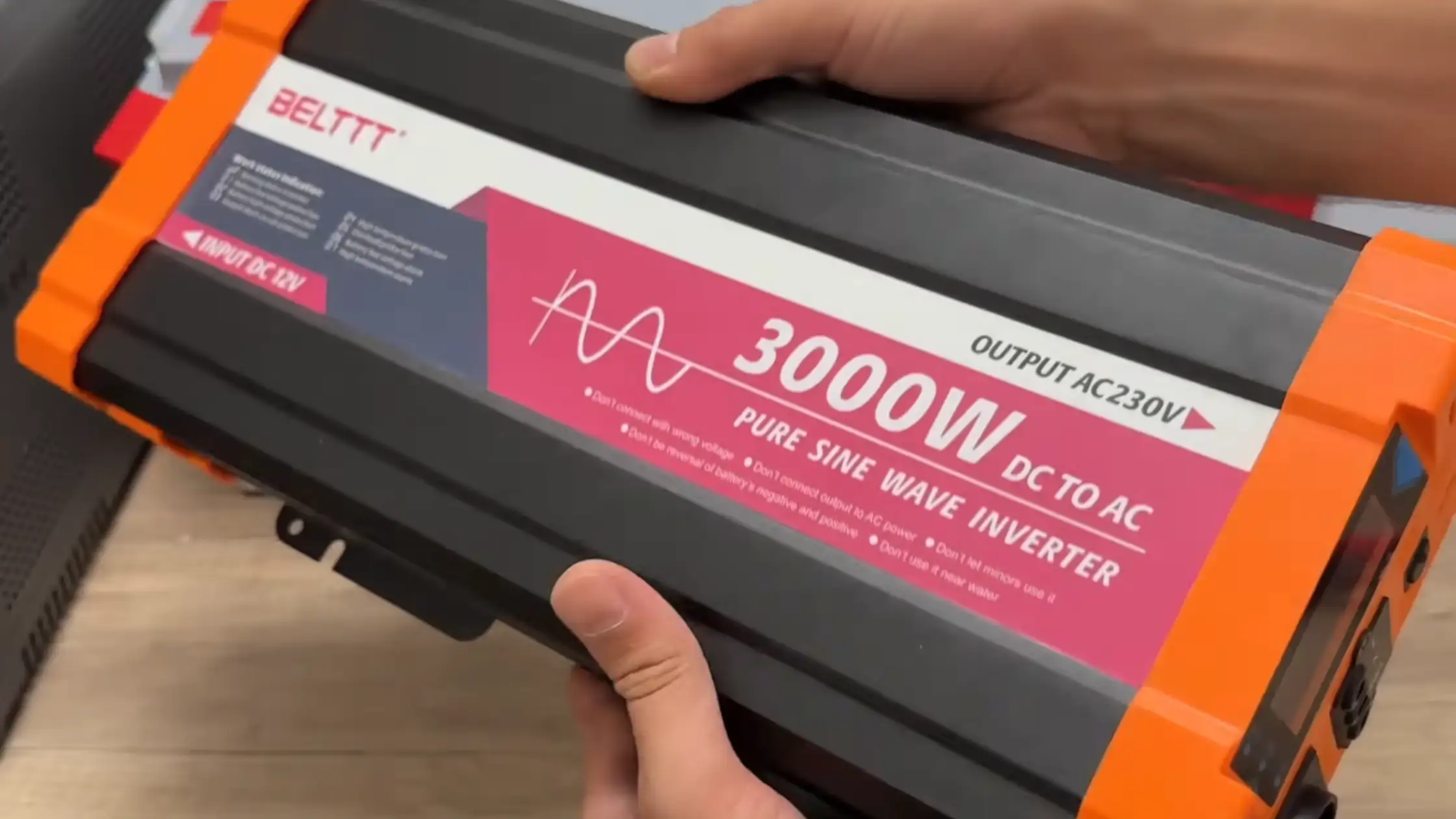 Close-up of James Ndungu holding the BELTTT 3000W Pure Sine Wave Power Inverter, showcasing its rugged black metal chassis, orange protective end caps, and clearly labeled front specifications against a light wood-grain background.